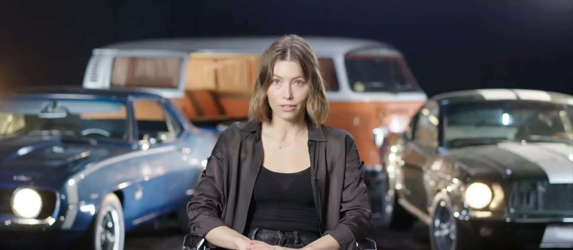 A cast member seated for an interview, with the orange and cream VW T2 camper van and two classic muscle cars parked behind her